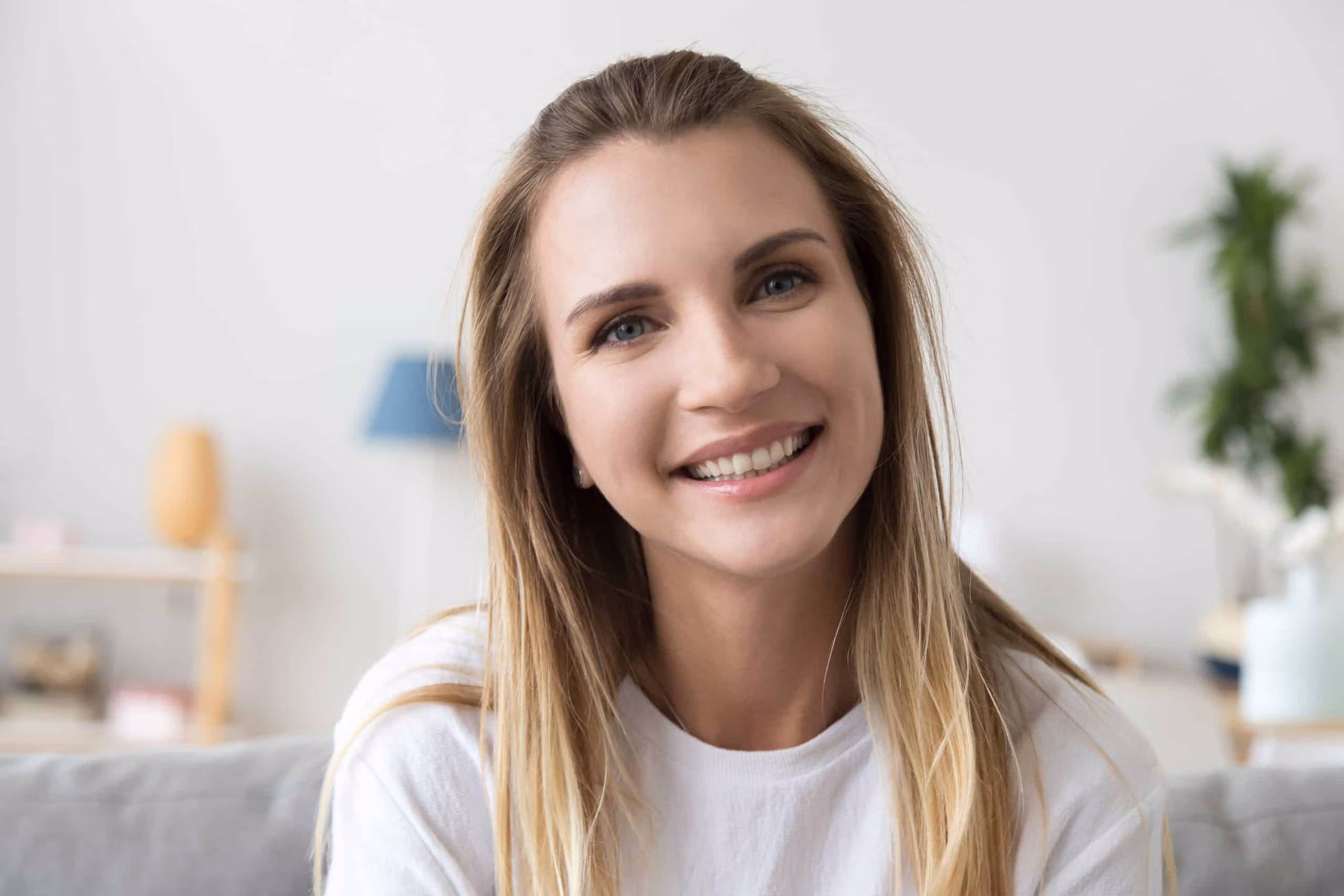 Support worker smiling indoors, seated in a home-like setting with a plant and lamp in the background.