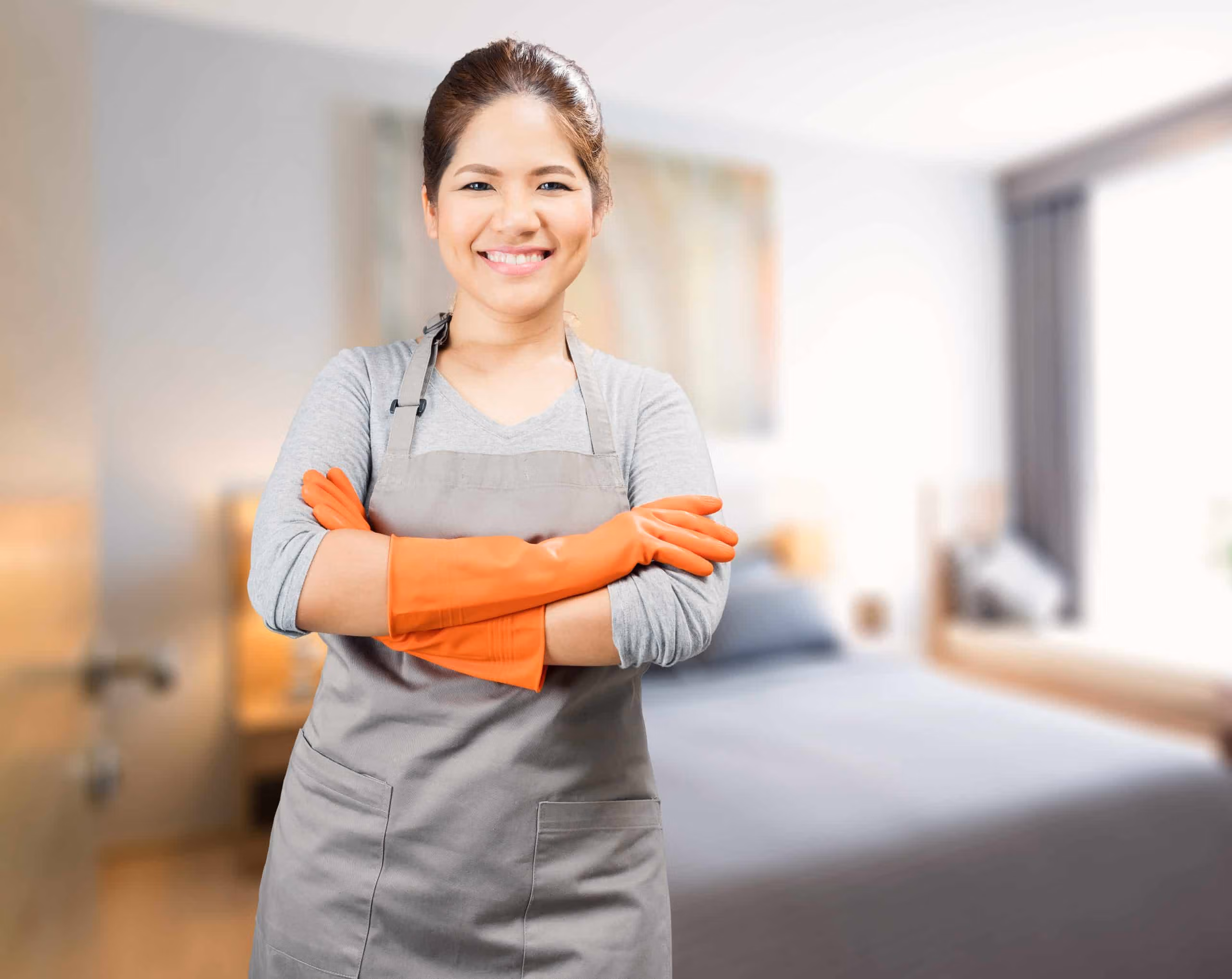 Support worker standing in a tidy room wearing an apron and bright orange kitchen gloves, smiling confidently.